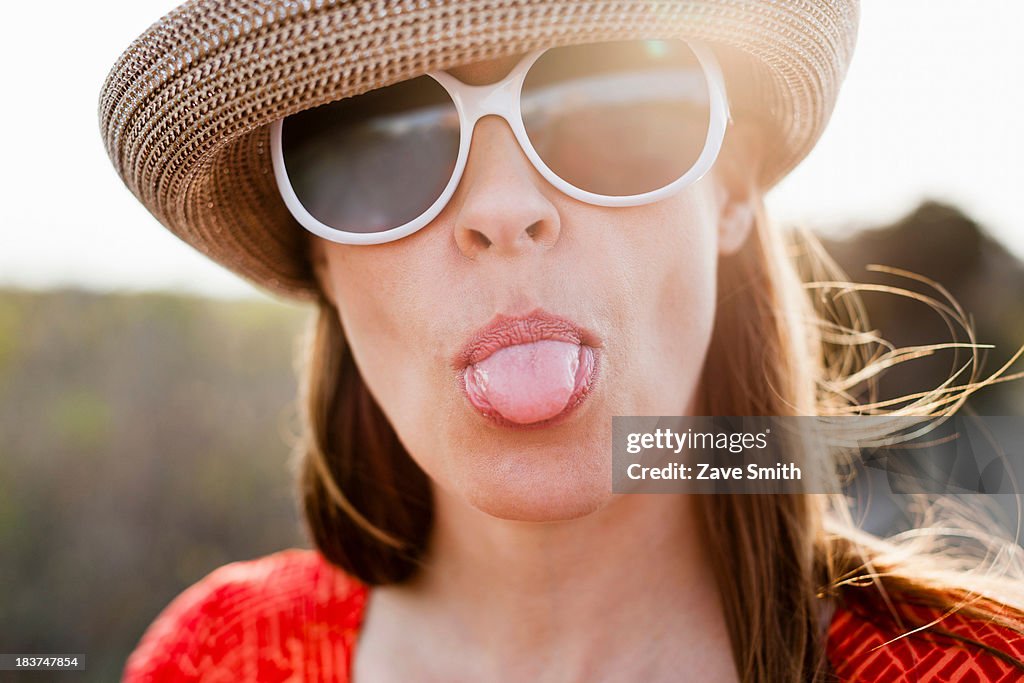 Mature woman wearing sunglasses and sunhat sticking out tongue
