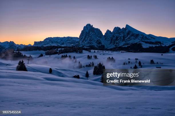 Hilly agricultural countryside with wooden huts and trees at Seiser Alm, Alpe di Siusi, covered in fresh snow in autumn at sunrise. The entire...