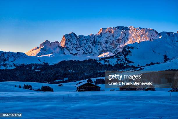 Hilly agricultural countryside with wooden huts and trees at Seiser Alm, Alpe di Siusi, covered in fresh snow in autumn at sunrise. The entire...