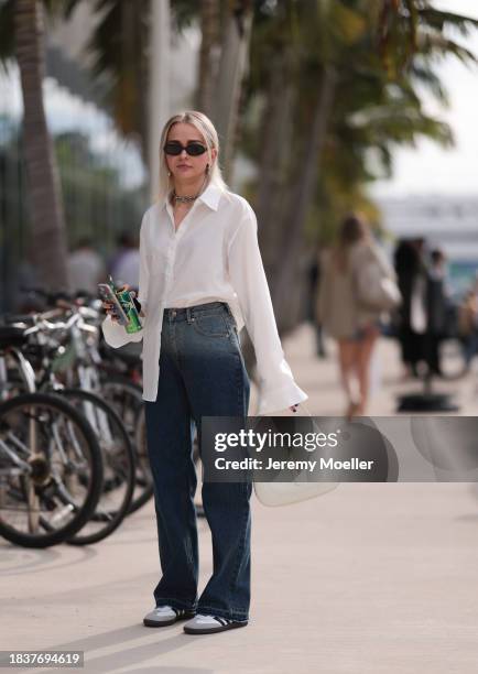 Olive Allen seen wearing black sunglasses, gold earrings and necklace, white buttoned shirt / blouse, light blue / dark blue faded wide leg denim...