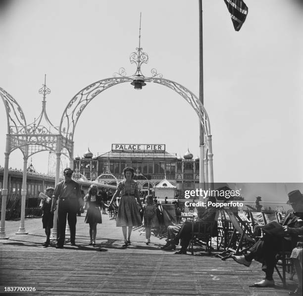 Family walks past visitors seated on deckchairs and directors chairs on the boardwalk of the Palace Pier in the seaside town of Brighton in Sussex,...