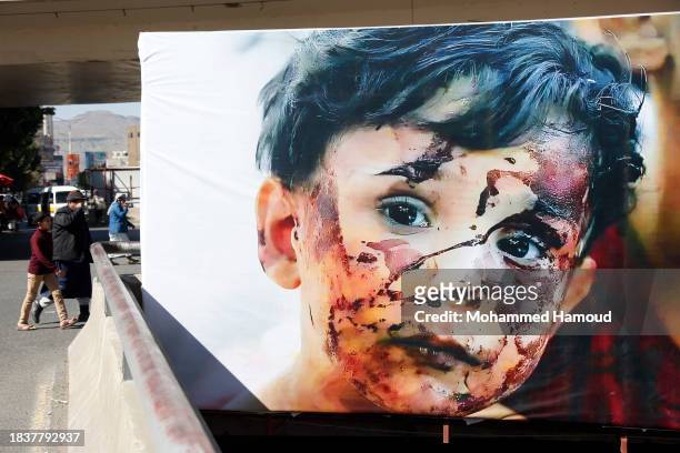 Yemeni passersby walk near a billboard bearing pictures of a Palestinian child with a blood-stained face after he was injured by an Israeli aerial...
