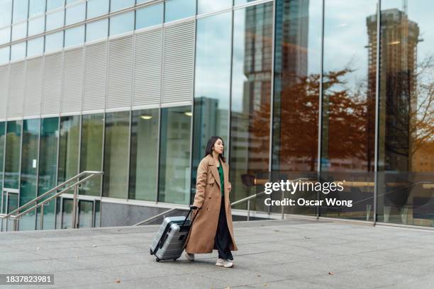 full length of young asian businesswoman carrying luggage, walking on city street against commercial buildings - business park uk fotografías e imágenes de stock