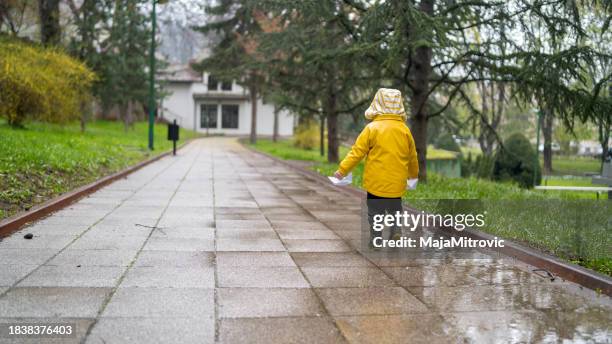 a small child in rubber boots and a jacket runs through puddles, has fun, plays and launches paper boats. - raincoat stock pictures, royalty-free photos & images