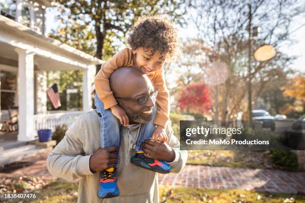 father carrying toddler boy on shoulders in front of home - schwarze schuhe stock-fotos und bilder