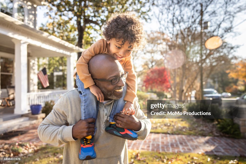 Father carrying toddler boy on shoulders in front of home