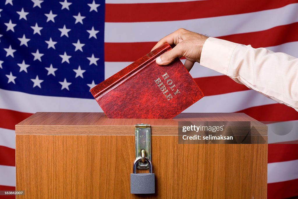 Human Hand Inserting Bible To Ballot Box Before American Flag