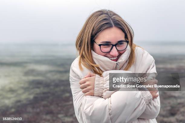 a smiling young woman with glasses and winter clothing crosses her arms to keep warm in the cold and foggy surroundings. - jan 19 stock pictures, royalty-free photos & images