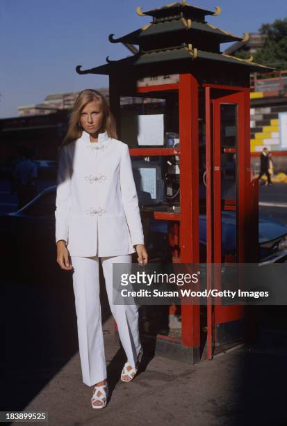 Portrait of an unidentified model dressed in a white, two-piece trouser suit as she poses beside a stylized phone booth, Manhattan's Chinatown, New...