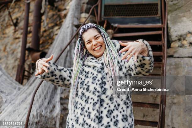 a positive charming girl with an unusual hairstyle. a laughing student in a fur coat with dreadlocks and african braids against the background of a ramshackle building. - student fashion stock pictures, royalty-free photos & images