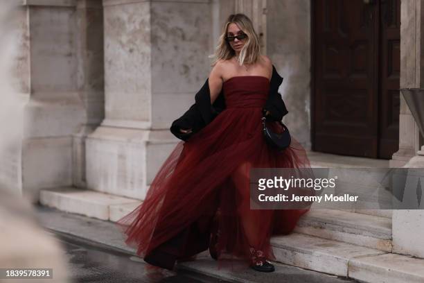 Karin Teigl seen wearing Saint Laurent black sunglasses, H&M Studio red ruffled tulle long bandeau dress, gold necklaces, H&M black wool long coat,...