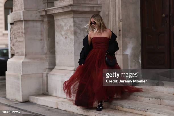 Karin Teigl seen wearing Saint Laurent black sunglasses, H&M Studio red ruffled tulle long bandeau dress, gold necklaces, H&M black wool long coat,...