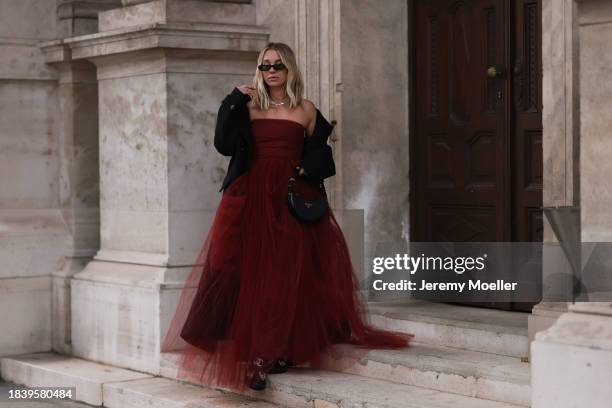 Karin Teigl seen wearing Saint Laurent black sunglasses, H&M Studio red ruffled tulle long bandeau dress, gold necklaces, H&M black wool long coat,...