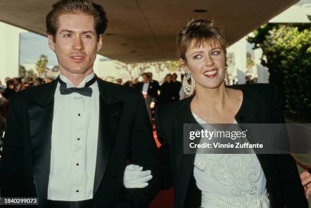 American actress Pamela Dawber, wearing a black jacket over a white outfit, walks arm-in-arm with her guest, who wears a tuxedo and bow tie, attends...