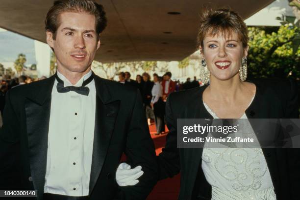 American actress Pamela Dawber, wearing a black jacket over a white outfit, walks arm-in-arm with her guest, who wears a tuxedo and bow tie, attends...