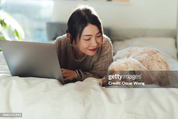 smiling young asian woman using laptop on bed with her dog lying next to her - pet -studio stock pictures, royalty-free photos & images