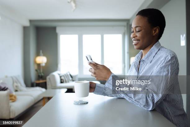 uma jovem mulher afro-americana com cabelo curto usando o telefone inteligente em casa e relaxando - women's short program - fotografias e filmes do acervo