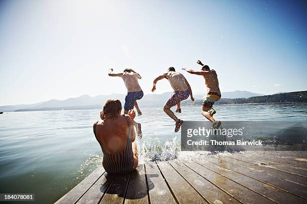 woman sitting on dock while men jump into water - floß stock-fotos und bilder