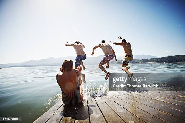 woman sitting on dock while men jump into water - group-of-friends-jumping-off-dock-into-lake stock pictures, royalty-free photos & images