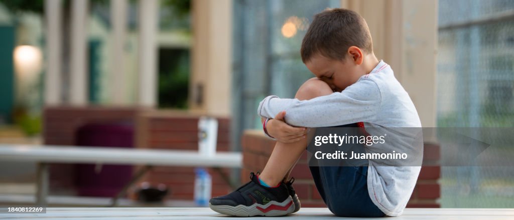Crestfallen Crying child boy sitting at the schoolyard covering his face