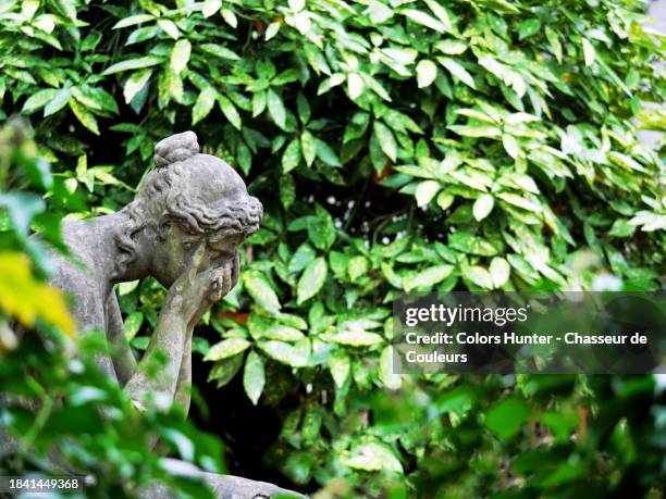 old stone sculpture depicting a sad woman, her face hidden in her hands and surrounded by bushes in paris, france - begraafplaats stockfoto's en -beelden