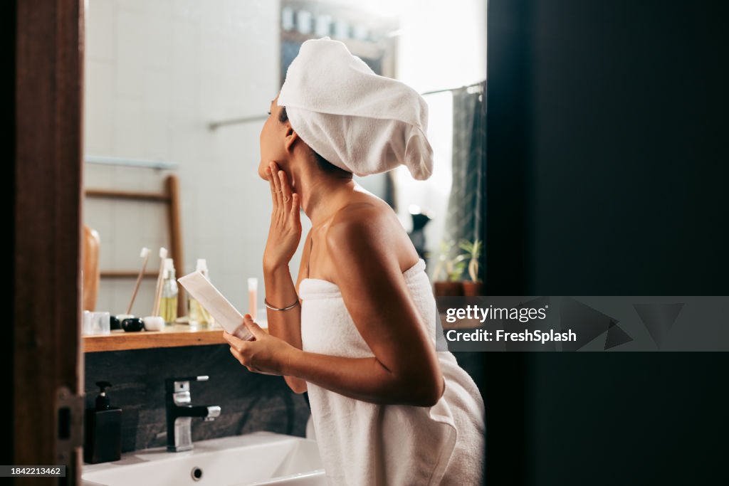 Una bella donna avvolta in un asciugamano che si guarda allo specchio mentre applica una crema sul viso dopo aver fatto il bagno