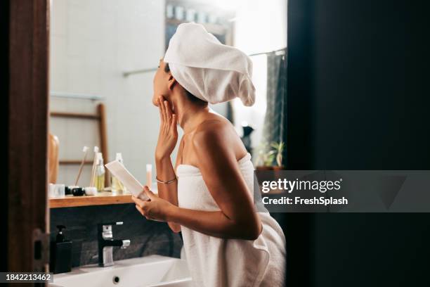 a beautiful woman wrapped in a towel looking herself in the mirror while applying some creme on her face after taking a bath - domestic bathroom stock pictures, royalty-free photos & images