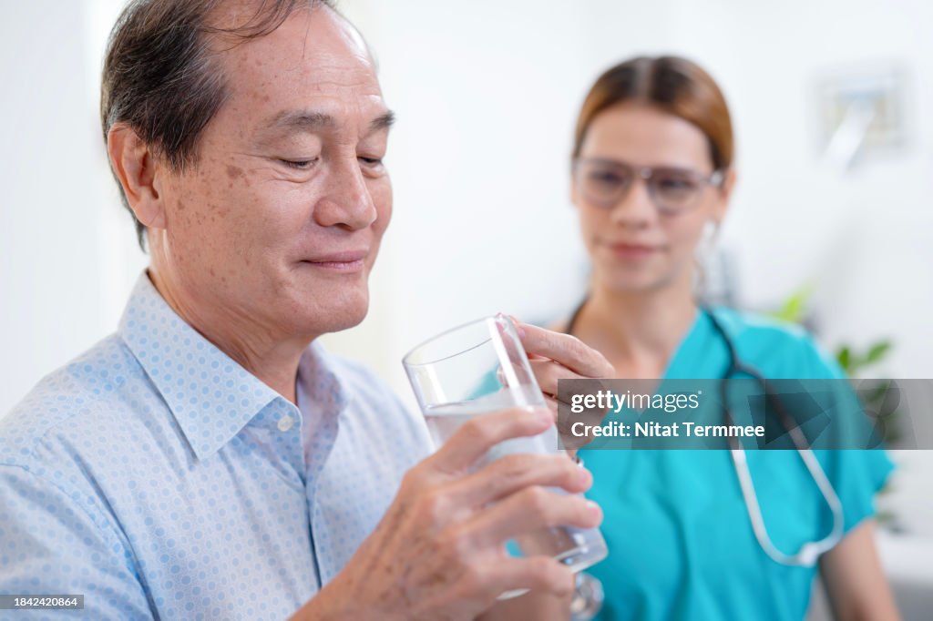 Taking Medicine Helps Control Blood Sugar Levels and Preven Diabetes in Geriatric. An Asian senior man taking pills with glass of fresh water while home nurse beside him to give advice.