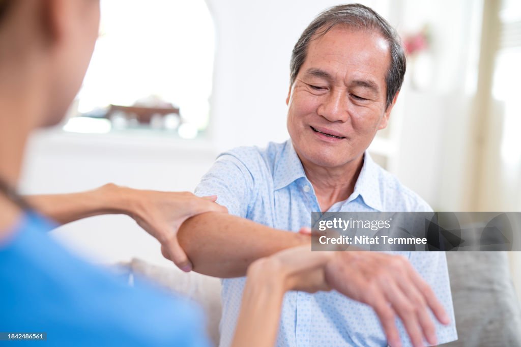 Geriatric Physical Therapy for Older Adults to Maintaining Independence in Performing Daily Activities. Home nurse helping a senior Asian man with massage of his hand and physical therapy to reduce myasthenia gravis disease at home.