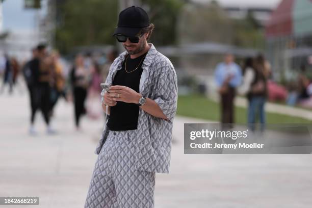 Eddy Piddington seen wearing black baseball cap, black sunglasses, gold necklace, black cotton t-shirt, Dior grey triangle pattern buttoned shirt,...