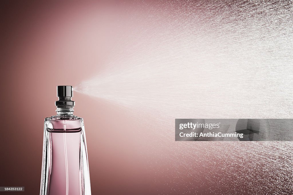 Glass bottle of perfume spraying mist against pink background