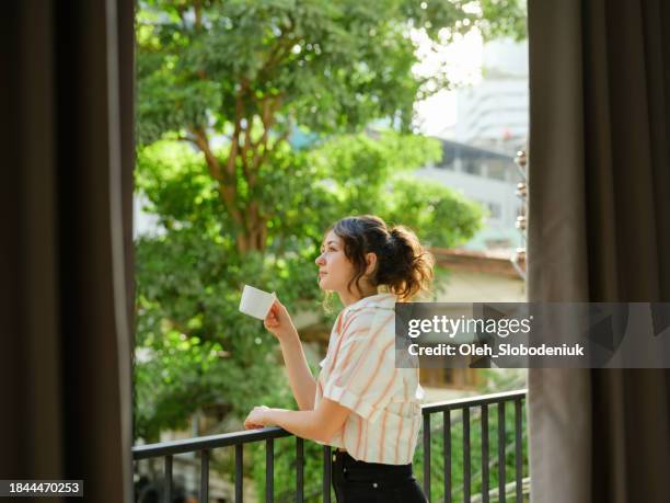 woman drinking coffee on the balcony in summer with trees visible from the window - balkong bildbanksfoton och bilder