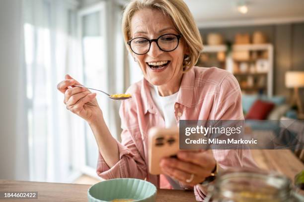 eine ältere frau isst cornflakes mit milch zum frühstück und sitzt im küchenbereich ihrer modernen wohnung - 50 60 jahre brille stock-fotos und bilder