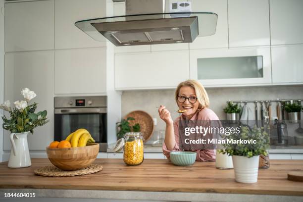 eine ältere frau isst cornflakes mit milch zum frühstück und sitzt im küchenbereich ihrer modernen wohnung - 50 60 jahre brille stock-fotos und bilder