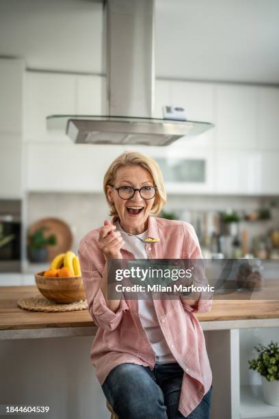 eine ältere frau isst cornflakes mit milch zum frühstück und sitzt im küchenbereich ihrer modernen wohnung - 50 60 jahre brille stock-fotos und bilder