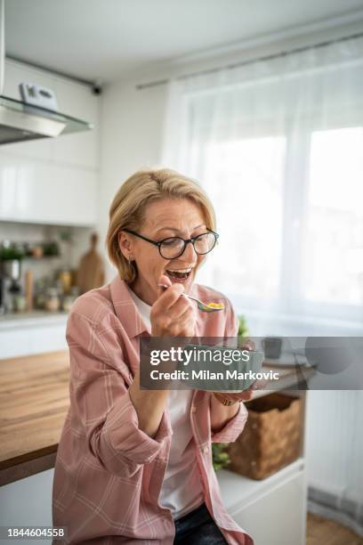 eine ältere frau isst cornflakes mit milch zum frühstück und sitzt im küchenbereich ihrer modernen wohnung - 50 60 jahre brille stock-fotos und bilder