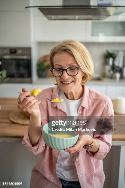 eine ältere frau isst cornflakes mit milch zum frühstück und sitzt im küchenbereich ihrer modernen wohnung - 50 60 jahre brille stock-fotos und bilder