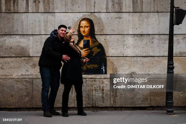 Couple takes selfies with their mobile phones of a street art poster, made by French artist Big Ben, depicting Leonardo da Vinci's Mona Lisa holding...