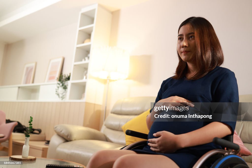 Donna incinta felice sulla sedia a rotelle con un piccolo sorriso seduta in salotto che tiene la pancia