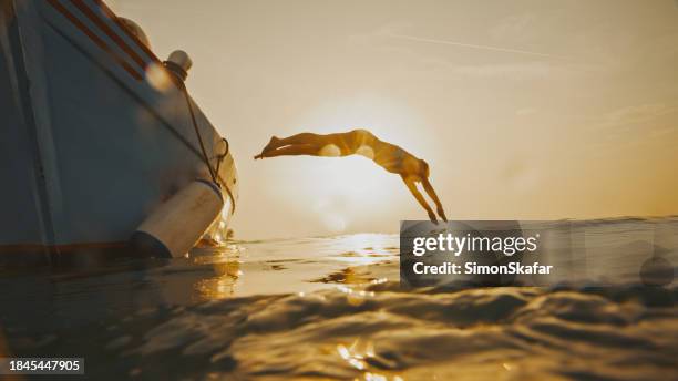 ganzkörper-seitenansicht einer von hinten beleuchteten frau, die während des sonnenuntergangs vom motorboot gegen den himmel ins meer taucht - taking the plunge stock-fotos und bilder