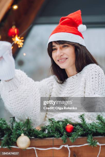 winter downtime with sparklers. woman in santa hat, waves a sparkler in christmas decorations outdoors. winter respite in a forest log cabin - bengalischer feuer stock-fotos und bilder