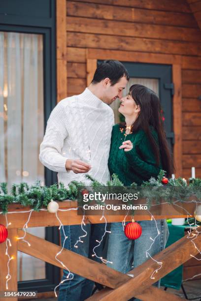 loving couple with sparklers, rubbing noses on wooden porch of log cabin on christmas eve - bengalischer feuer stock-fotos und bilder