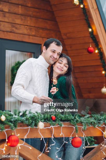 christmas glamping vacation for two: hugging romantic couple sparks off sparklers on the porch of a log cabin on vacation - bengalischer feuer stock-fotos und bilder