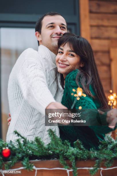 couple winter cheer with sparklers: woman and man hug and rejoice on the deck of a wooden house during winter - bengalischer feuer stock-fotos und bilder