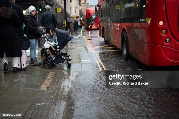 As a London bus splashes past, a woman with a child in its pushchair waits to cross the road but looks worried at the puddle on Piccadilly on a rainy...