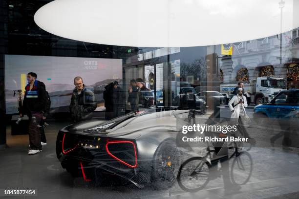 Reflections of a cyclist, pedestrians and potential buyers looking at a Lotus sports car which is displayed in the window of the brand's showroom on...