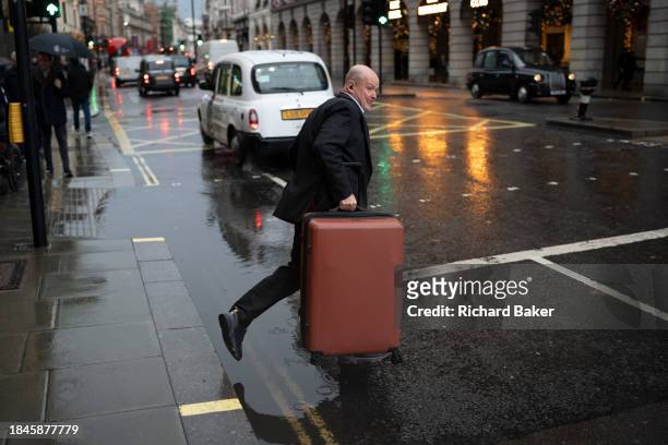 Man with a suitcase finds a gap in the traffic to cross Piccadilly on a rainy afternoon in Westminster, on 12th December 2023, in London, England.