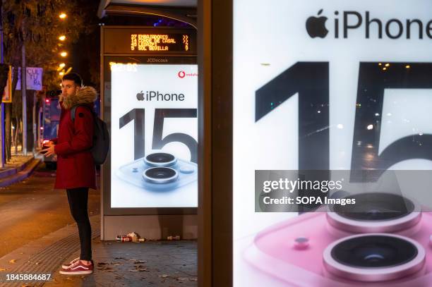 Commuter waits at a bus stop next to an American multinational technology company Apple iPhone 15 commercial advertisement in Spain.