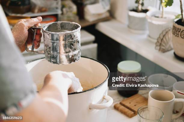 grind the flour in an iron calico mug over a baking sheet with dough. atmospheric photo of homemade baked goods on the kitchen table - powdered sugar sifter fotografías e imágenes de stock