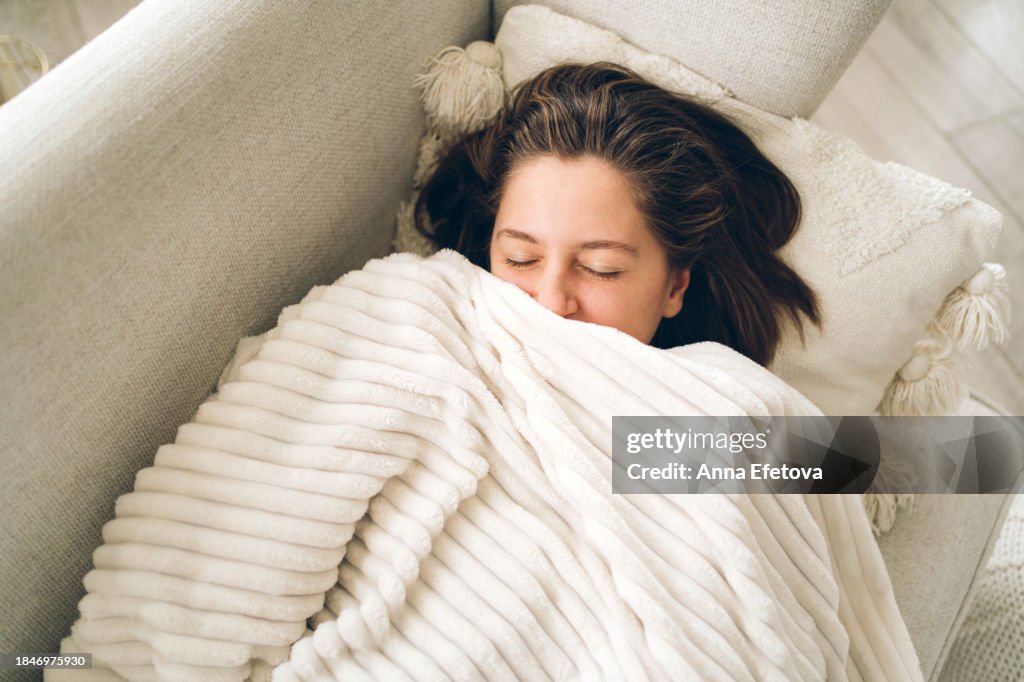 Young smiling woman lies on the couch and looks out from under the blanket. She has gray eyes and short brown hair. Photography from above